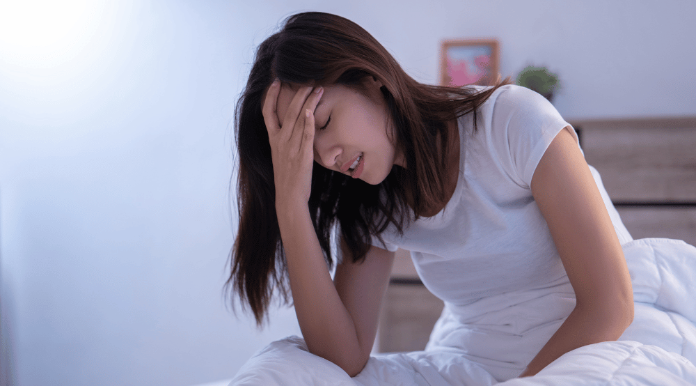 Woman sitting on her bed and holding a hand to her head in pain