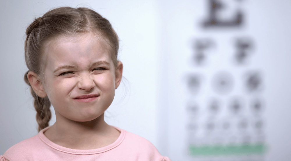 a little girl squinting with a blurred eye chart in the background